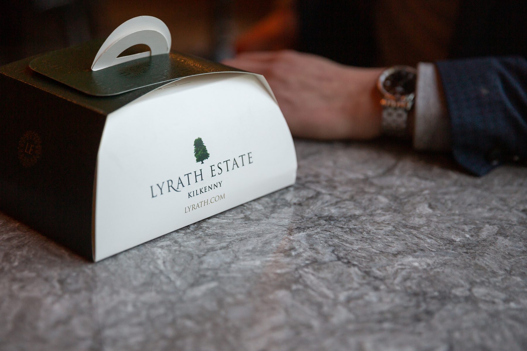 Dark green and white patisserie box with a curved top and white plastic carry handle. Printed with 'LYRATH ESTATE KILKENNY' logo and small tree graphic.