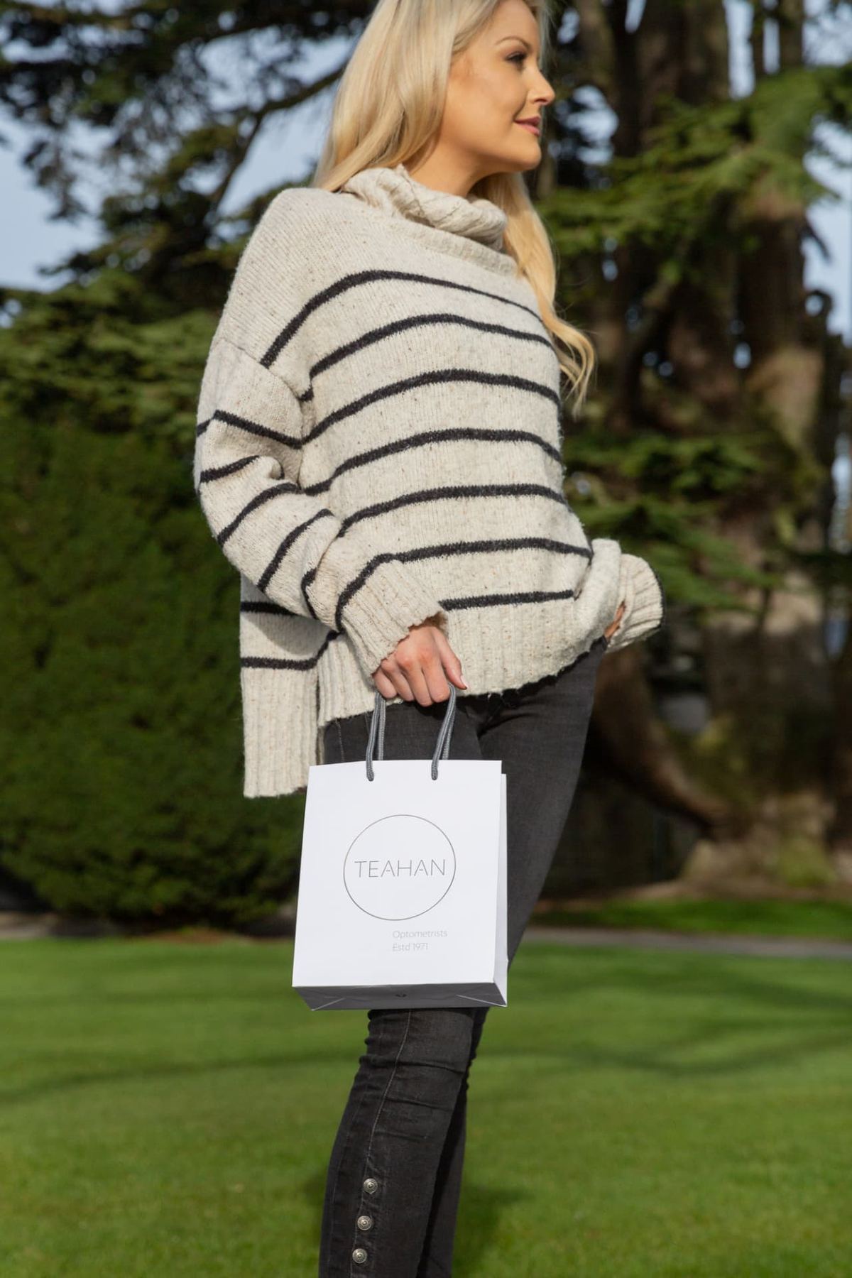 Small white matt laminated portrait paper bag with grey rope handles and a circular grey printed logo for 'TEAHAN Optometrists & Opticians'.