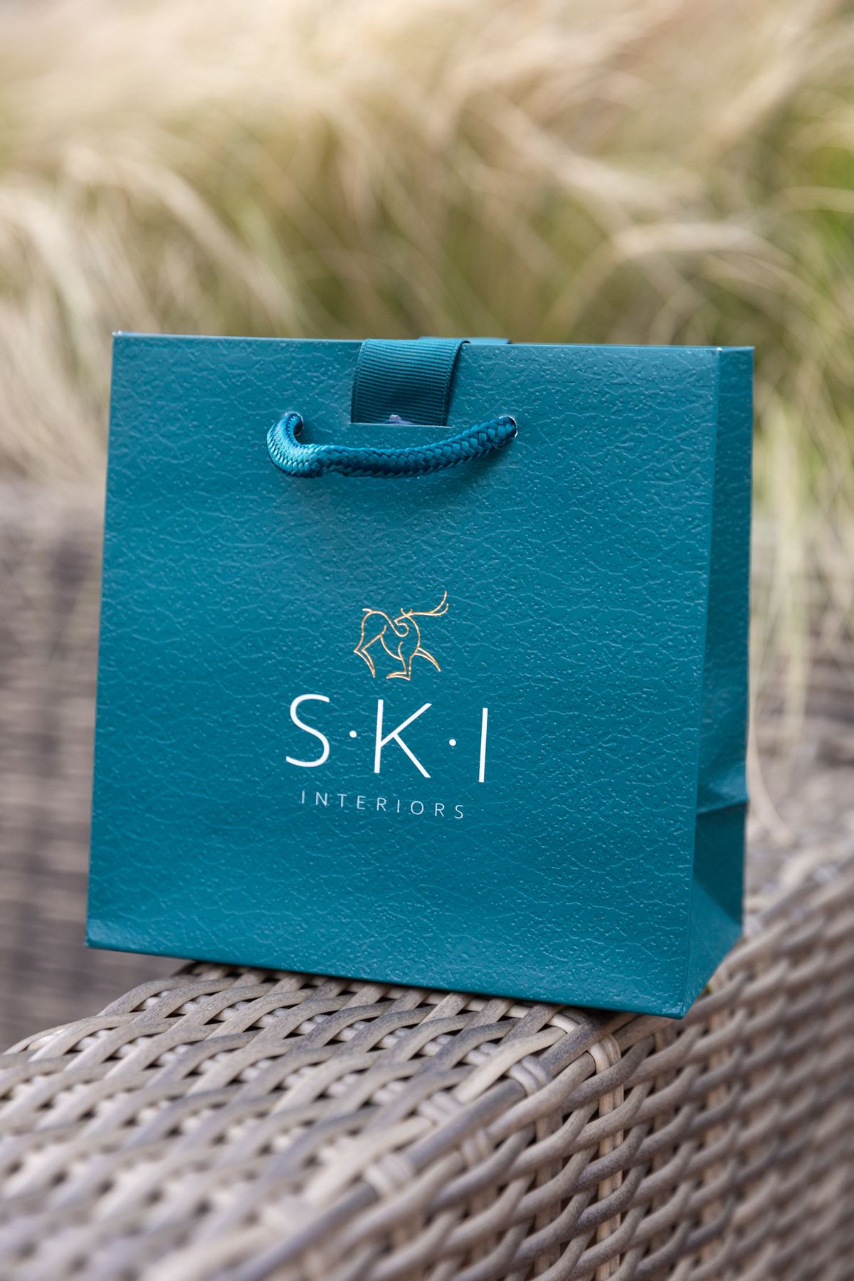 Small square teal paper gift bag with an embossed textured finish, gold foil logo, and matching teal braided rope handles with ribbon reinforcement.