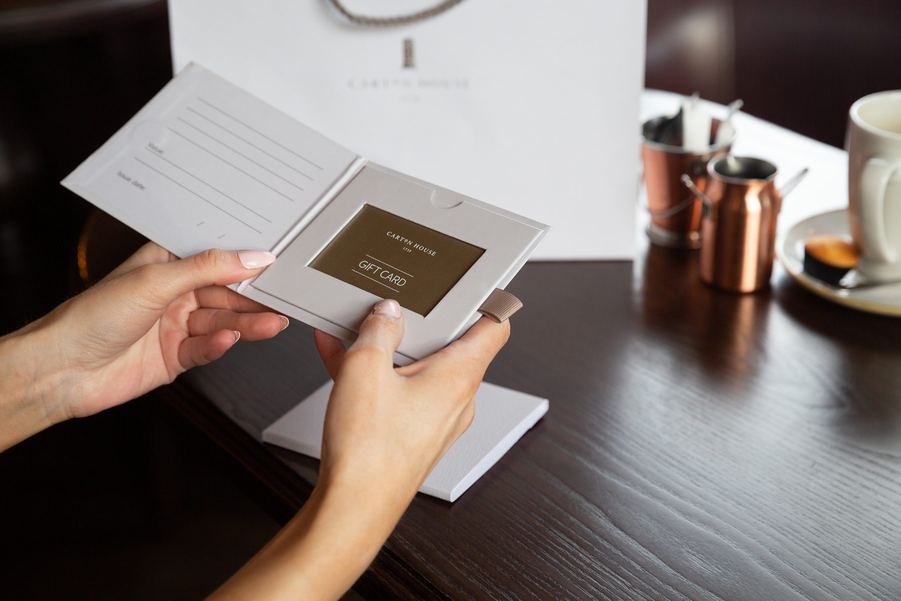 Light grey rigid gift card wallet, gold foil logo, ribbed pull tab, internal card slot. A light grey paper bag is in the background.