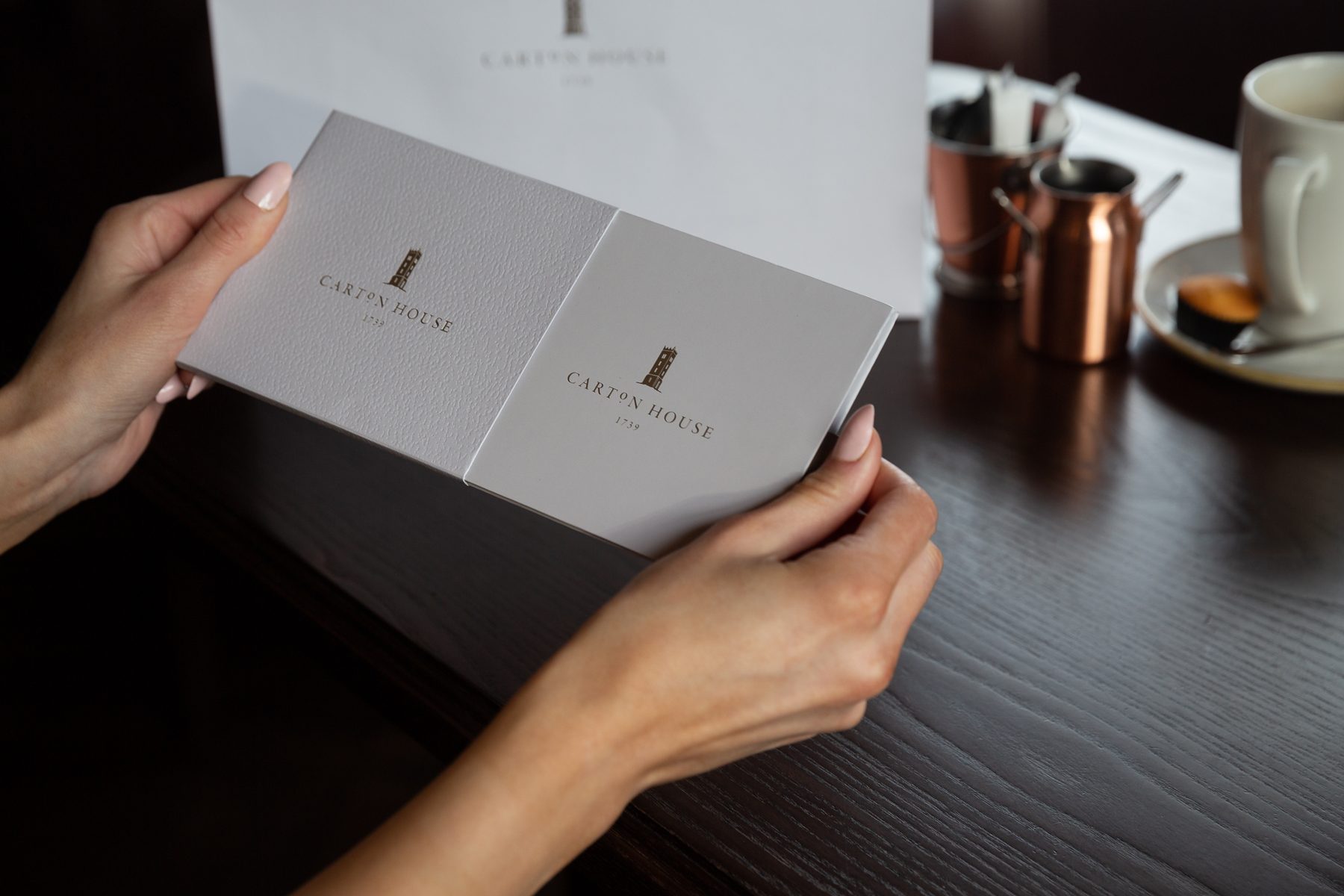 A light grey rigid box, textured finish, with a gold foil "CARTON HOUSE" logo. Next to it, a matching white paper carrier bag.
