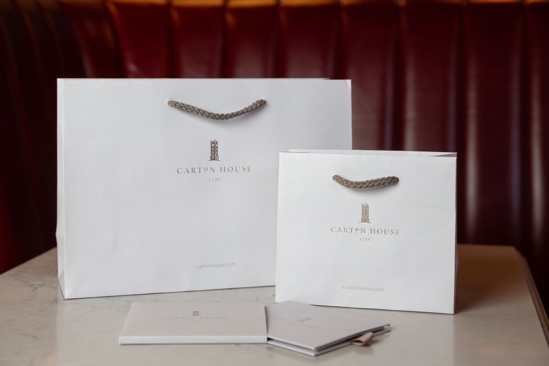 Two white matt laminated paper bags of different sizes, with dark beige braided rope handles and a subtle light brown printed logo and website.