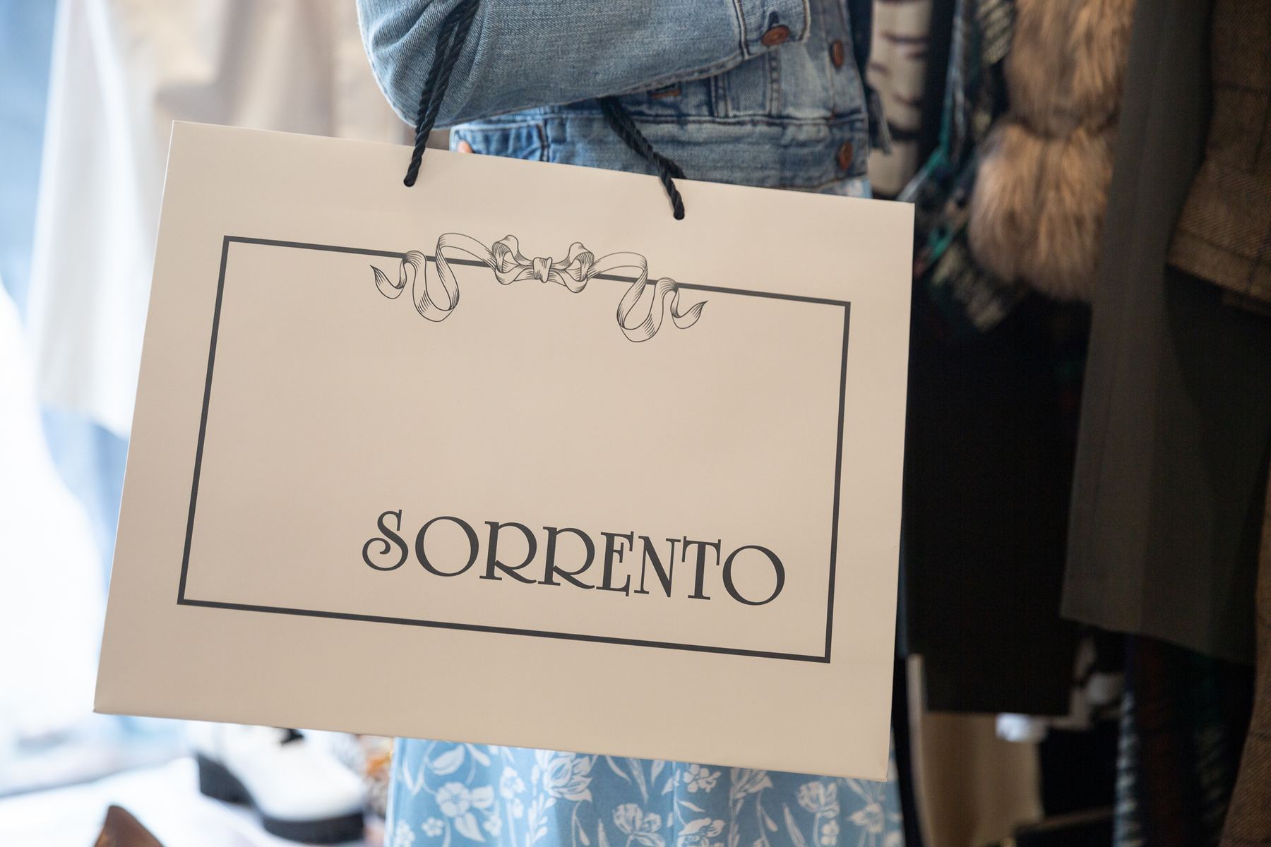 Landscape cream paper bag with black rope handles. Features a black printed border, decorative ribbon graphic, and 'SORRENTO' logo.