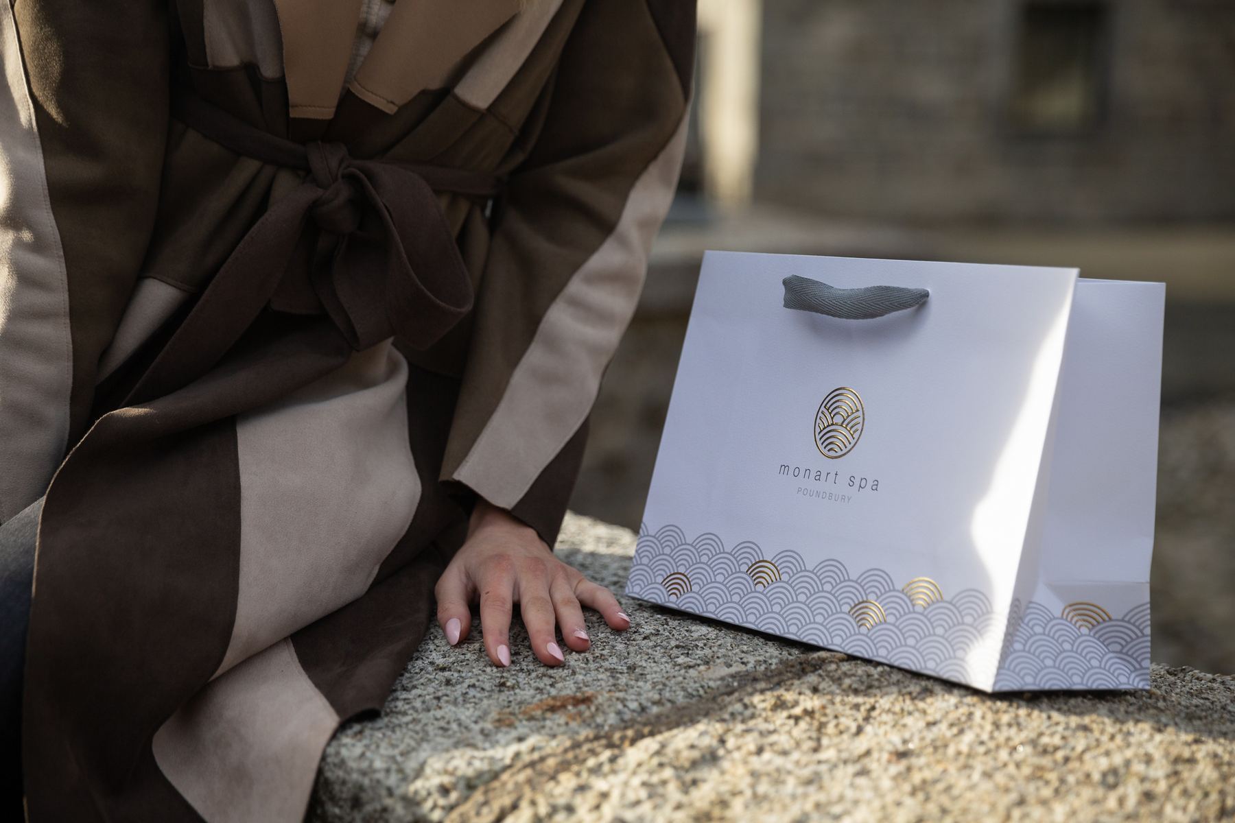Matt white paper carrier bag with gold and silver foil logo and wave pattern, featuring grey grosgrain ribbon handles.