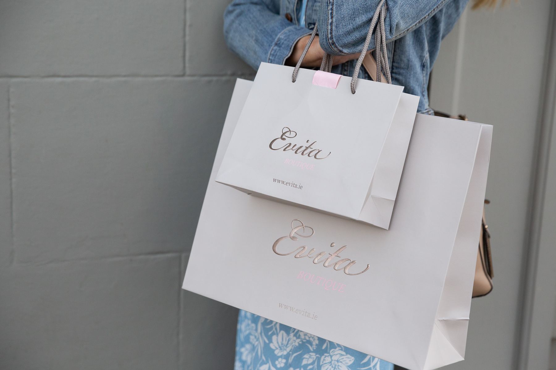 Two matt laminated light grey paper bags with rose gold foil logos, text, and grey rope handles. The smaller bag has a pink ribbon tab.