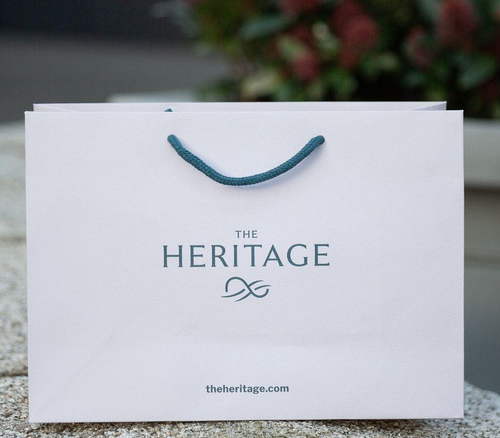 White landscape paper bag with blue green rope handles and "THE HERITAGE" logo in blue green print, landscape format.