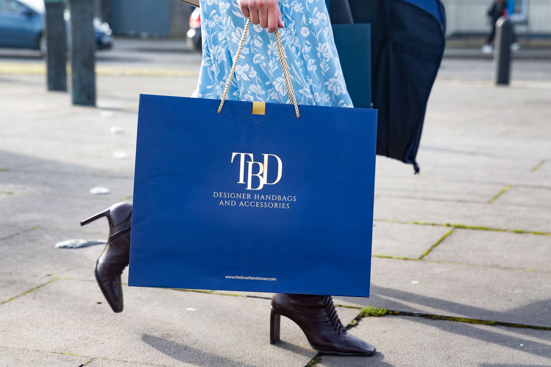 A landscape format matt navy paper carrier bag with a white foil logo and gold twisted rope handles, reinforced with gold ribbon tabs.