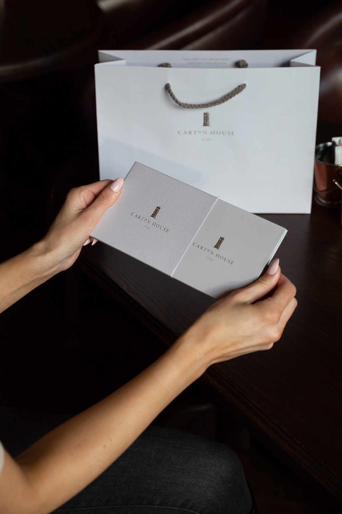 White landscape paper bag with brown rope handles and subtle gold foil logo. Matching grey gift card wallet, gold foil logo, debossed finish.