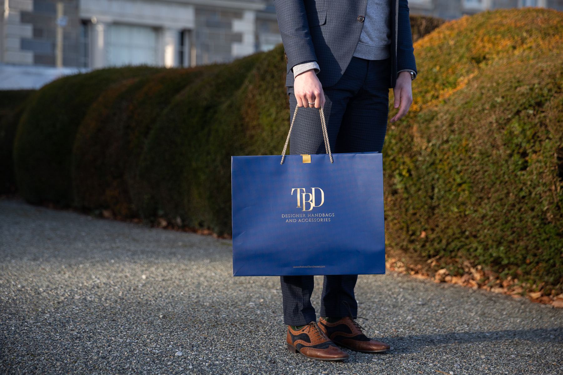 Navy blue matt laminated landscape paper carrier bag with gold foil TBD logo, gold twisted rope handles, and a gold ribbon tab closure.