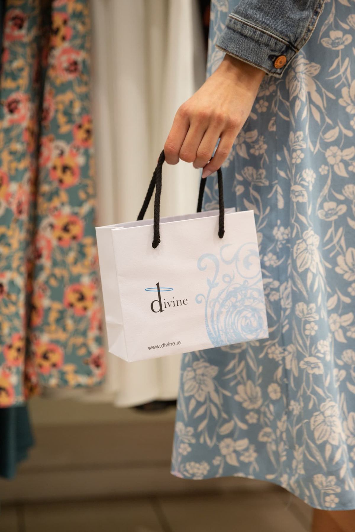 Small white paper carrier bag with black twisted rope handles, featuring a light blue and black 'Divine' logo with decorative swirls.