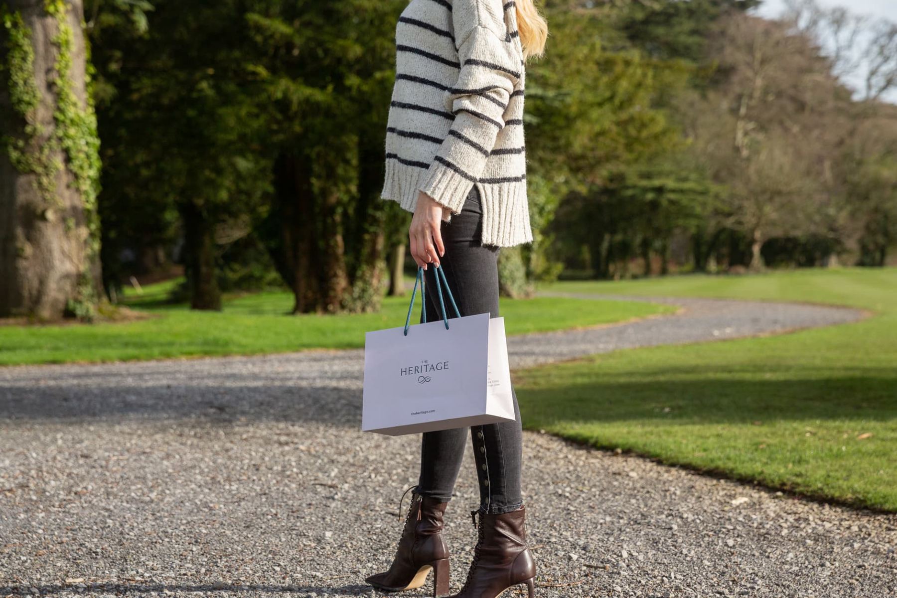 White matt laminated paper bag with 'The Heritage' logo in grey, featuring teal twisted rope handles, presented in a portrait orientation.