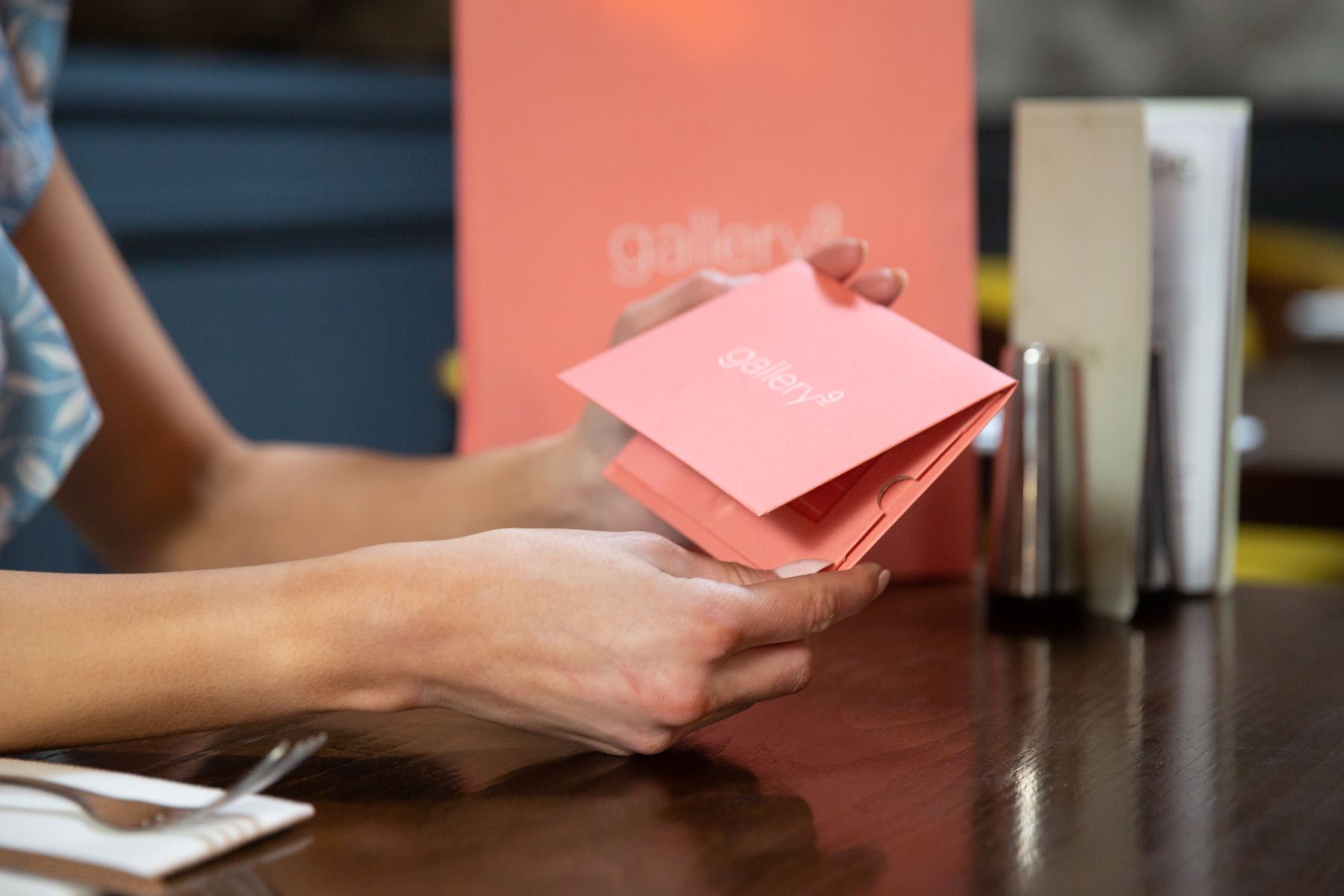 Coral folded card jewellery pouch with white 'gallery' logo. A small tab closure secures contents. A matching coral paper bag is in the background.