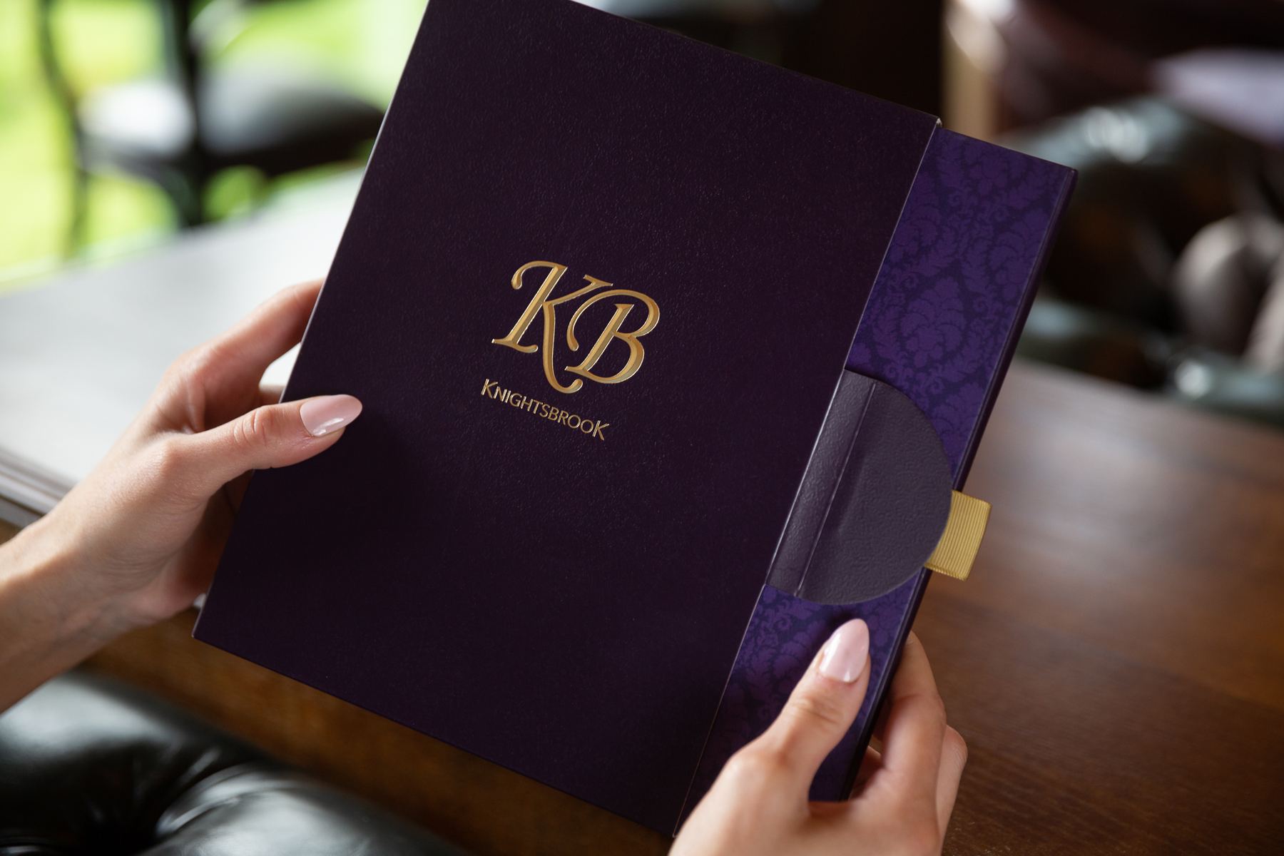 Purple rigid presentation box with gold foil logo, magnetic flap closure, and a gold ribbon tab. The side panel features a subtle patterned print.