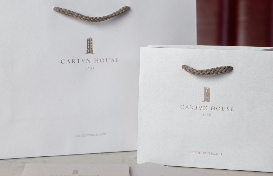 Two white matt laminated paper bags of different sizes, with dark beige braided rope handles and a subtle light brown printed logo and website.