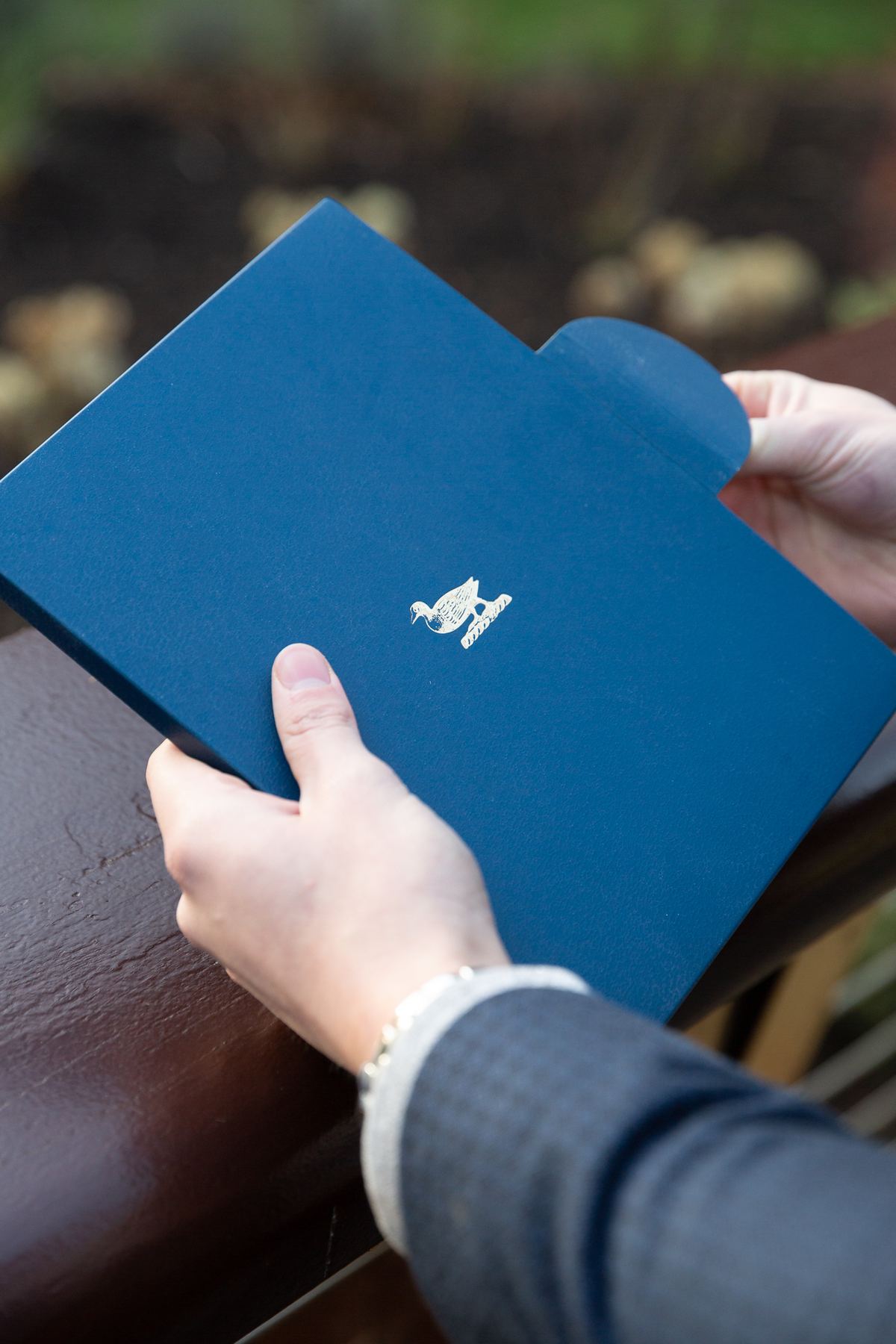 Navy blue card folder with a matt finish and a gold foil stamped logo featuring a duck and quill, with an integrated tab closure.