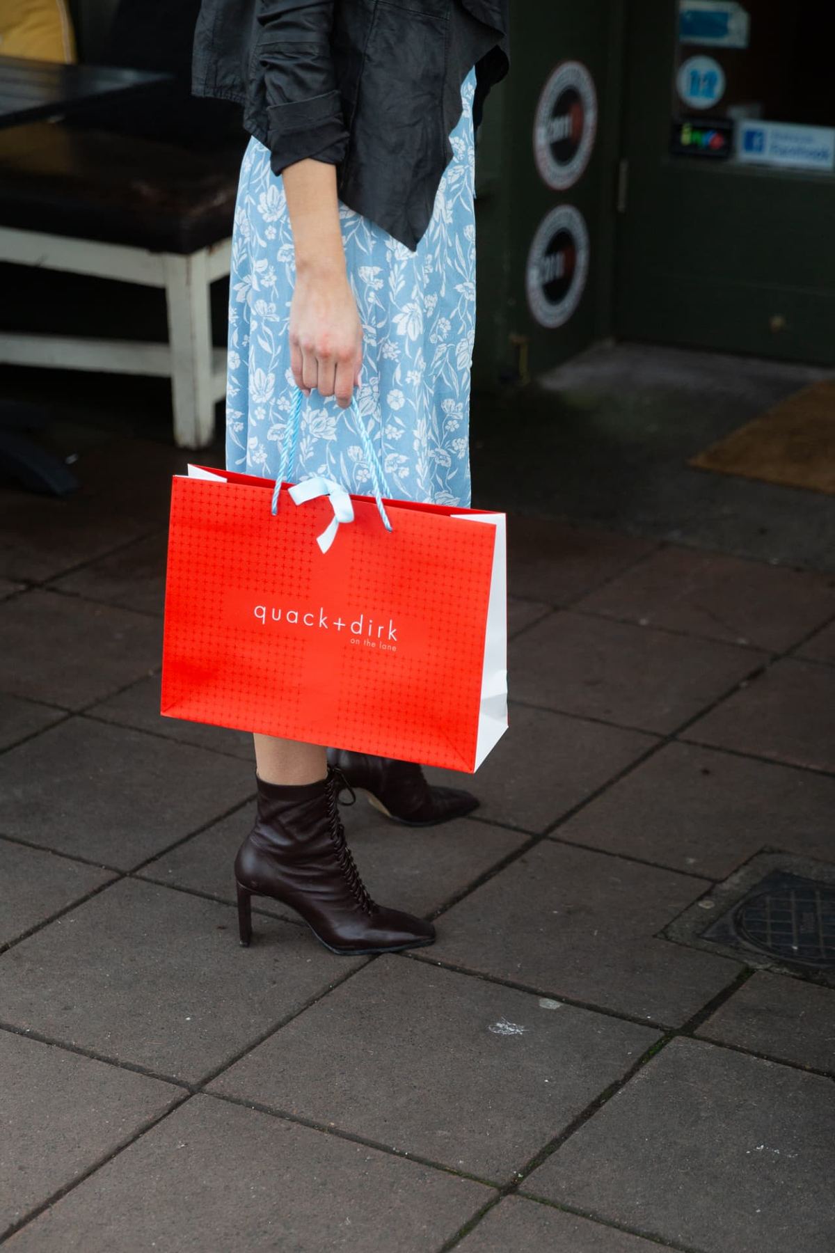 Red landscape paper carrier bag with white patterned background, white printed logo and light blue twisted rope handles, tied with white ribbon.