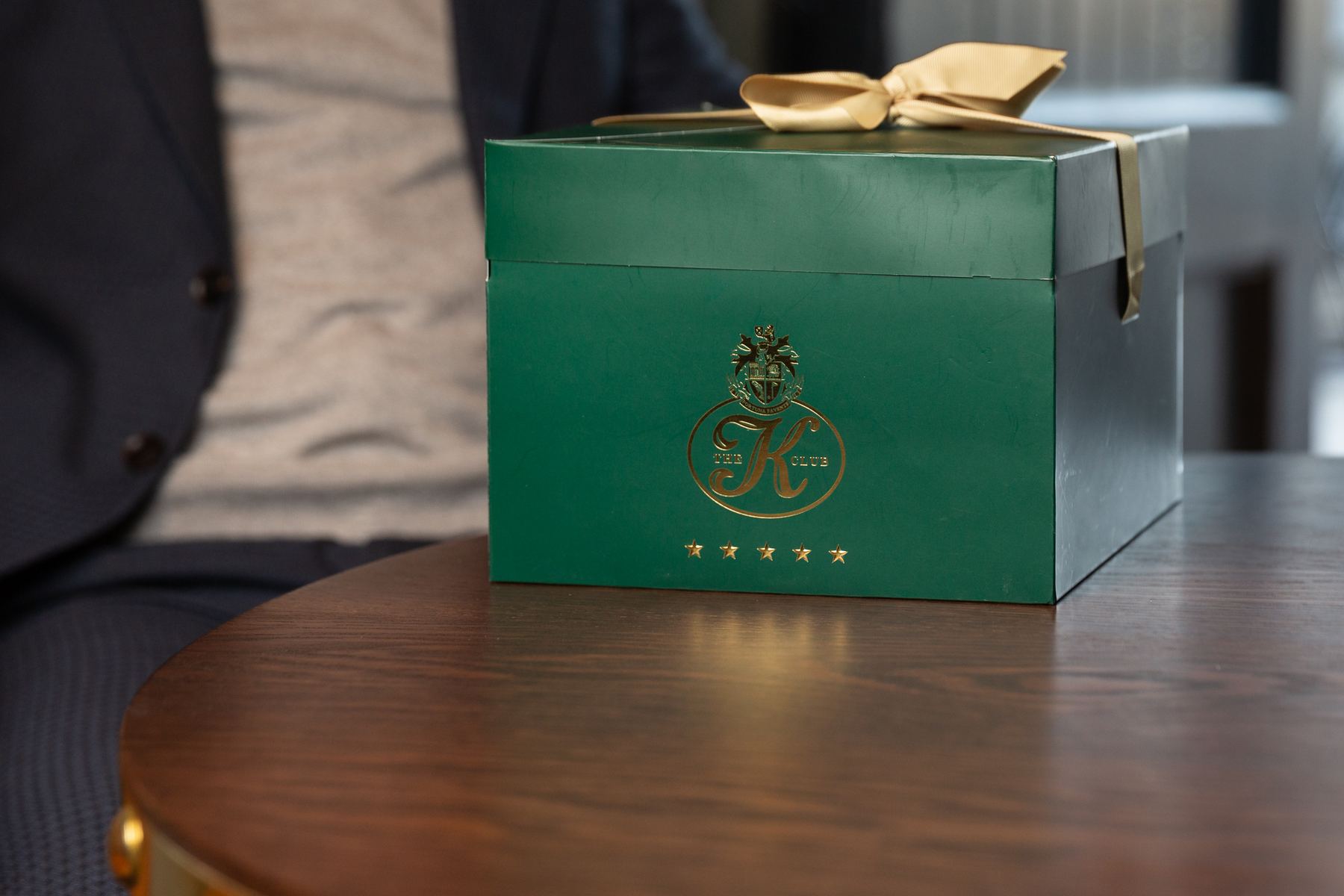 Deep green rigid gift box with gold foil logo and five stars, adorned with a decorative gold ribbon bow tied on the top lid.