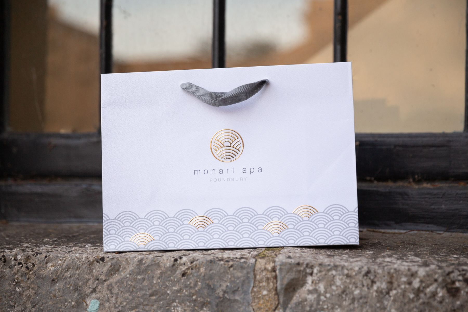 White matt laminated landscape carrier bag, grey ribbon handles, gold foil Monart Spa logo, grey gold wave pattern along bottom.