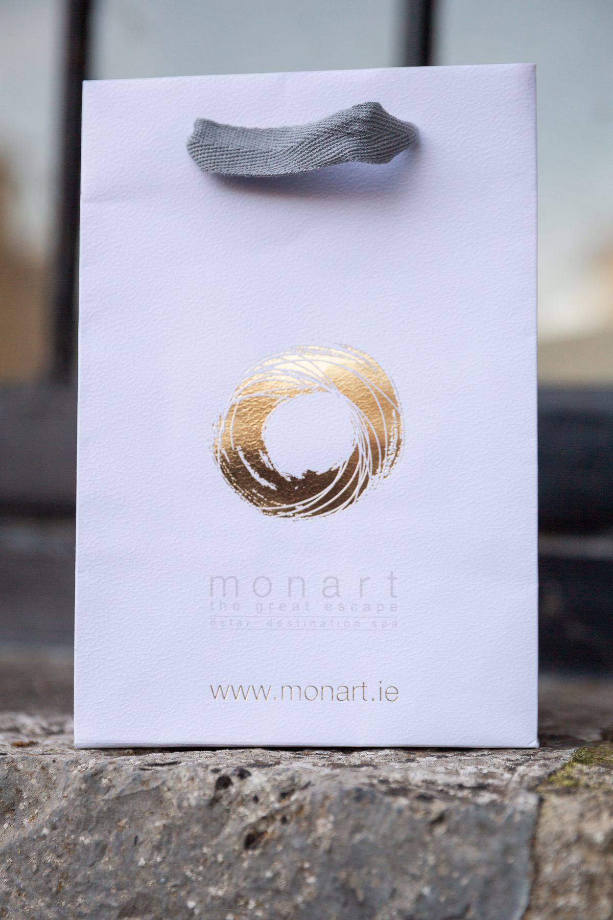 White portrait paper bag with an unlaminated textured finish, gold foil printed brushstroke logo, and grey grosgrain ribbon handle.