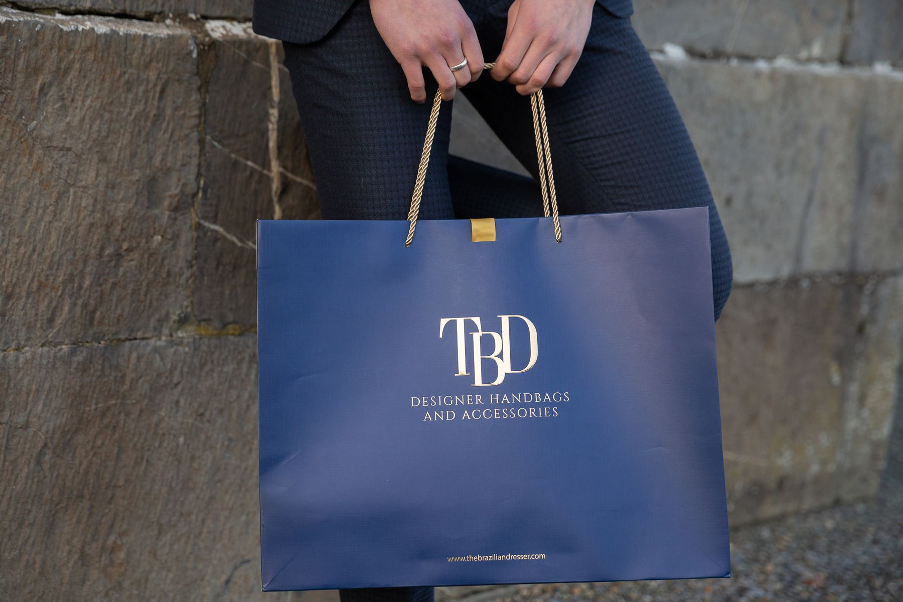 Navy blue matt laminated landscape paper bag with gold foil logo, gold rope handles, and a gold internal ribbon reinforcement for the handles.