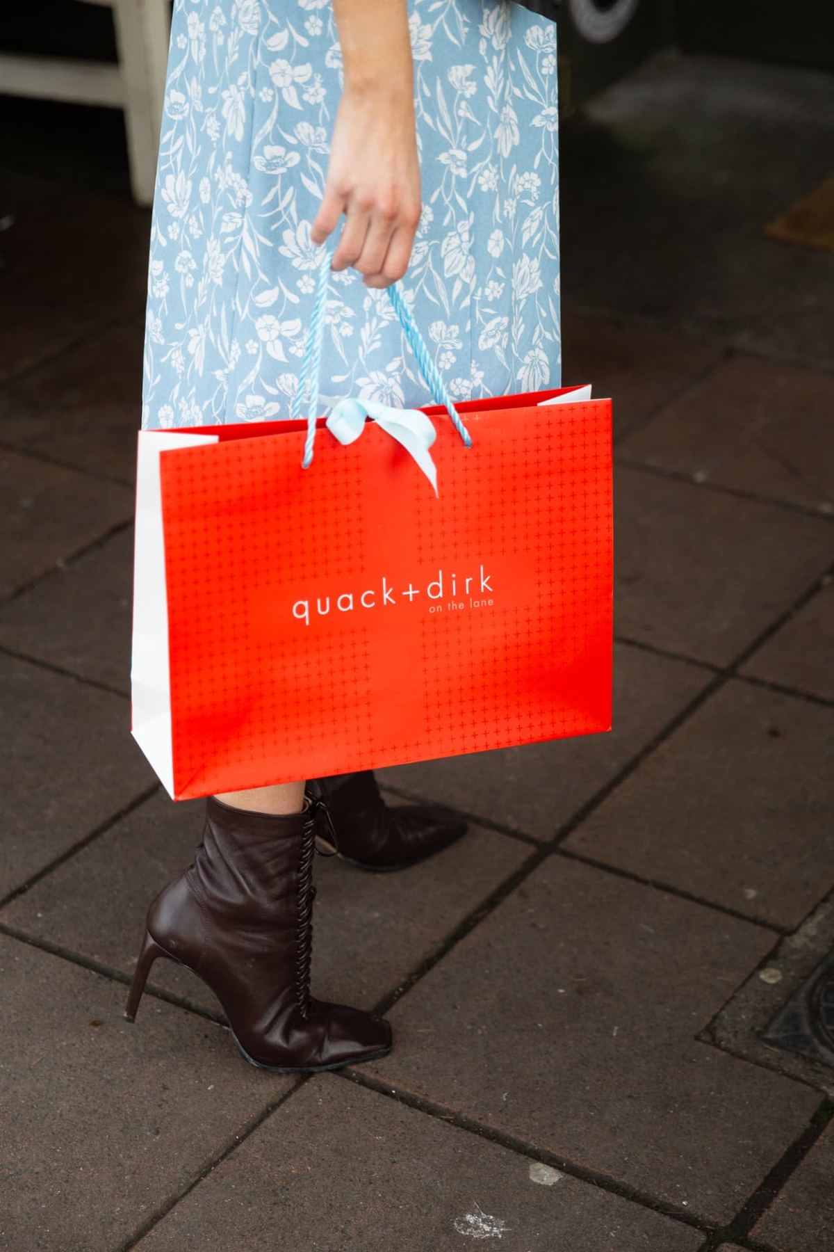 Red landscape paper bag with white gussets, white grid print, and white 'quack+dirk' logo. Light blue rope handles with a light blue ribbon bow.