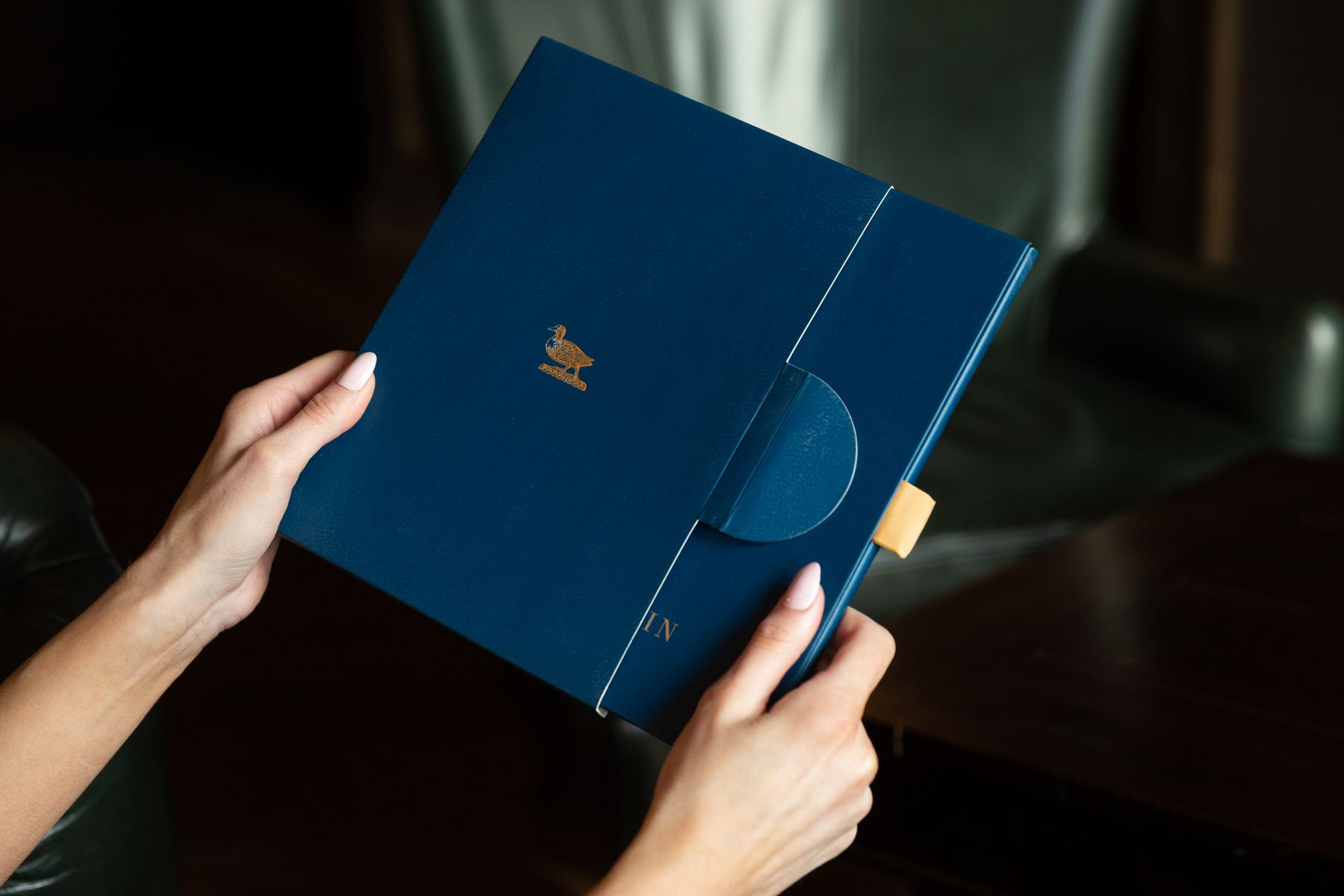 Navy matt rigid presentation box with gold foil duck logo, magnetic flap closure, and a beige fabric tab for opening.