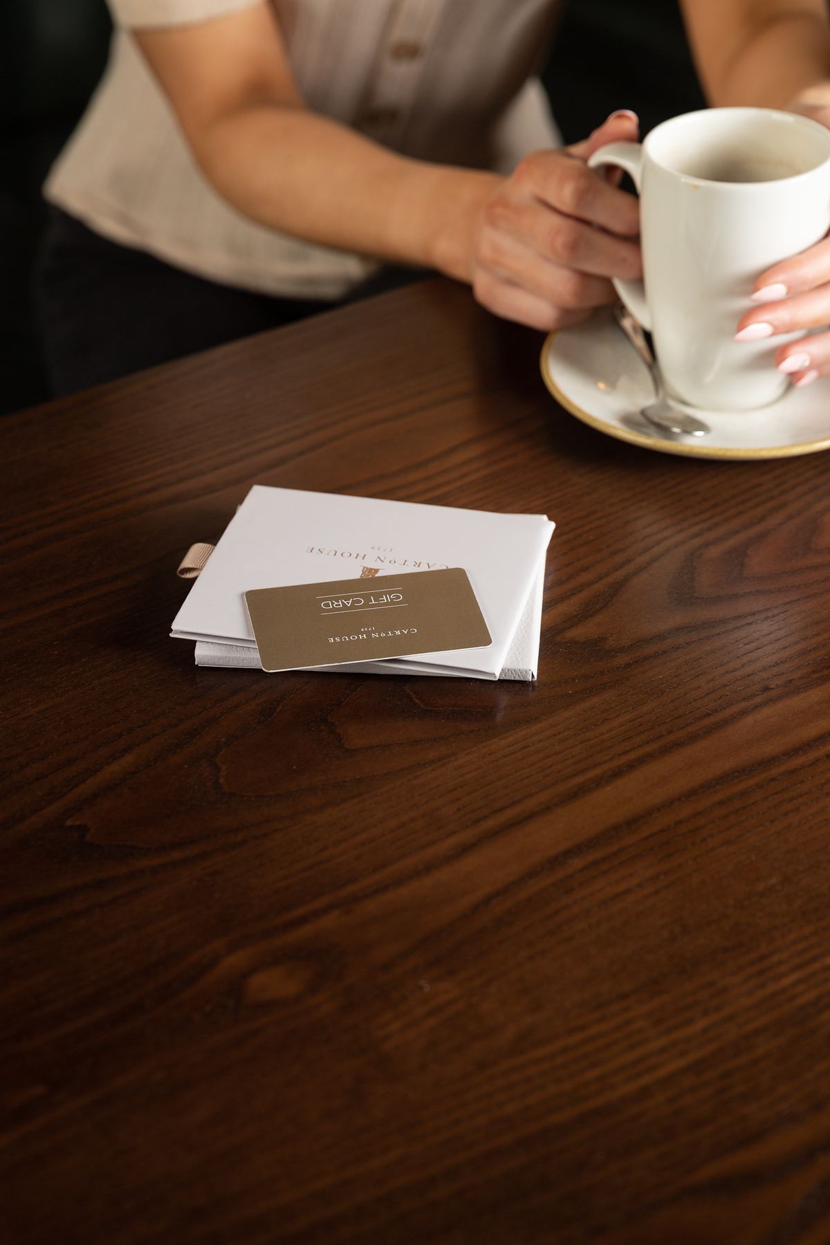White paper wallet with gold foil logo, housing a gold plastic gift card with black text. Features a beige ribbon pull tab.