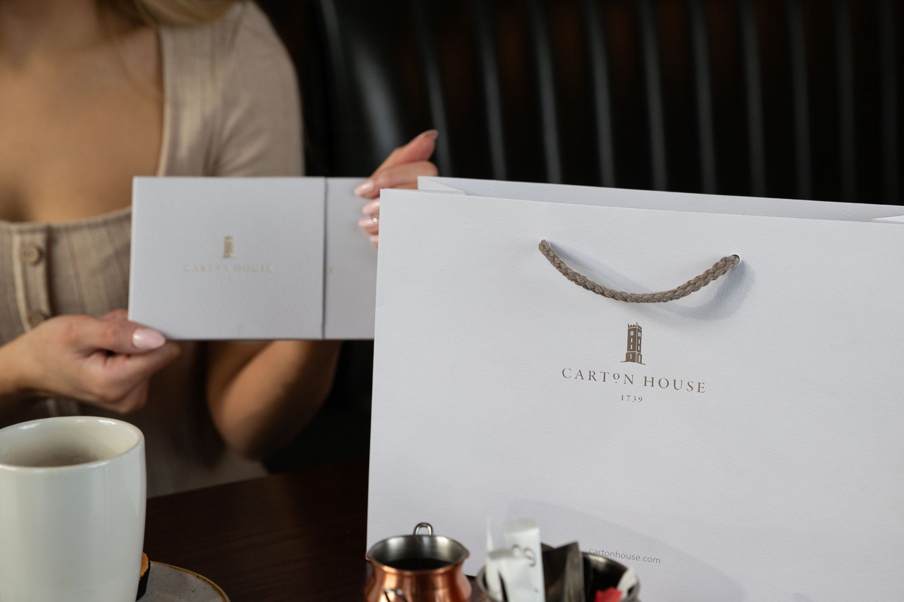White matt laminated paper bag with braided natural rope handles and a matching white gift card wallet with gold foil logo, luxury retail packaging.