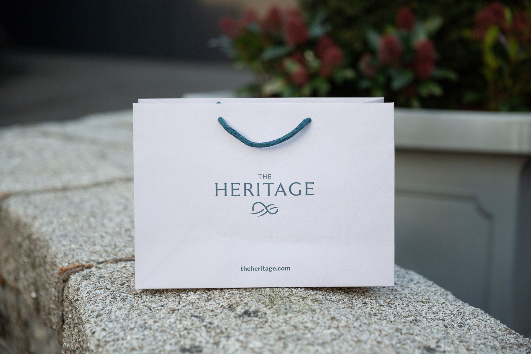 White landscape paper bag with blue green rope handles and "THE HERITAGE" logo in blue green print, landscape format.