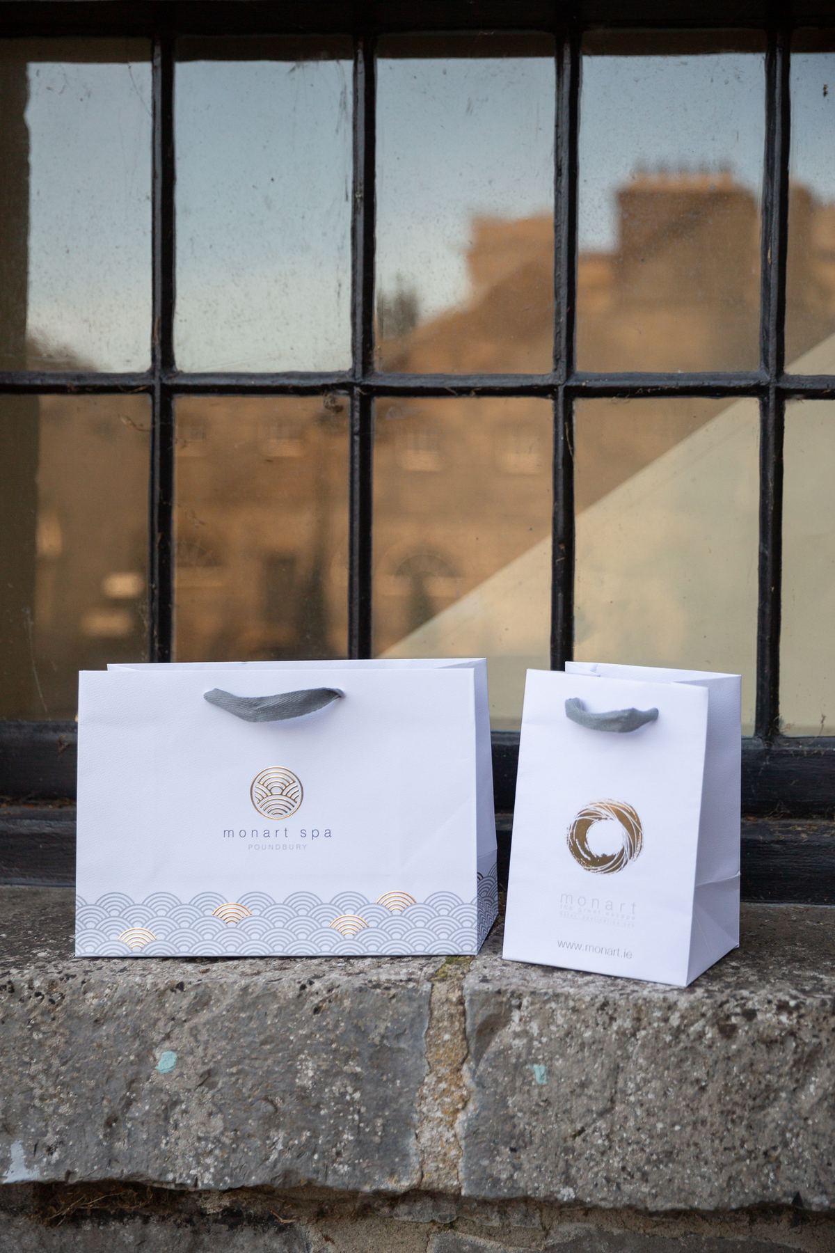 Two white matt laminated paper bags with grey ribbon handles and gold and grey foil print, one landscape, one portrait.