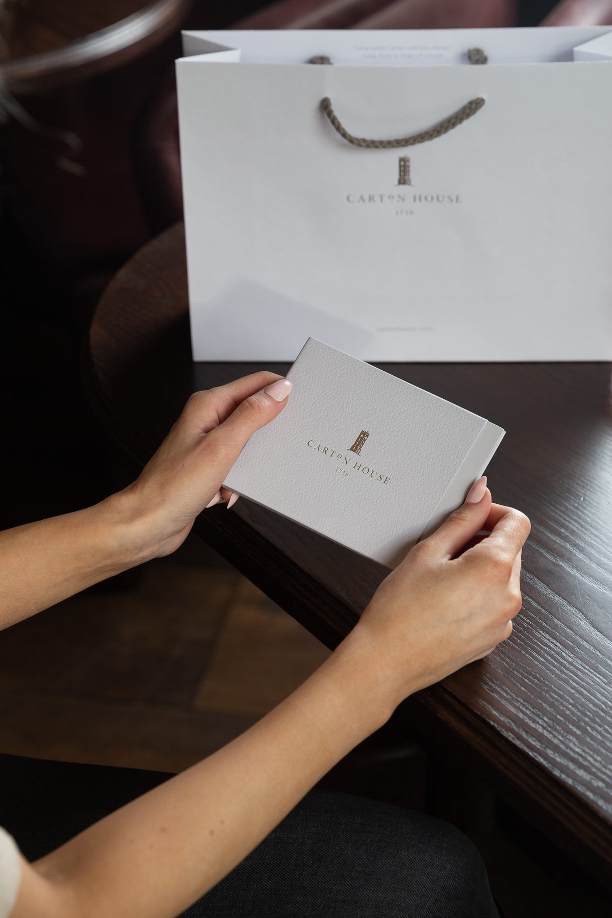 Light grey textured rigid gift box with gold foil logo and a white paper bag with a gold foil logo and grey rope handles.