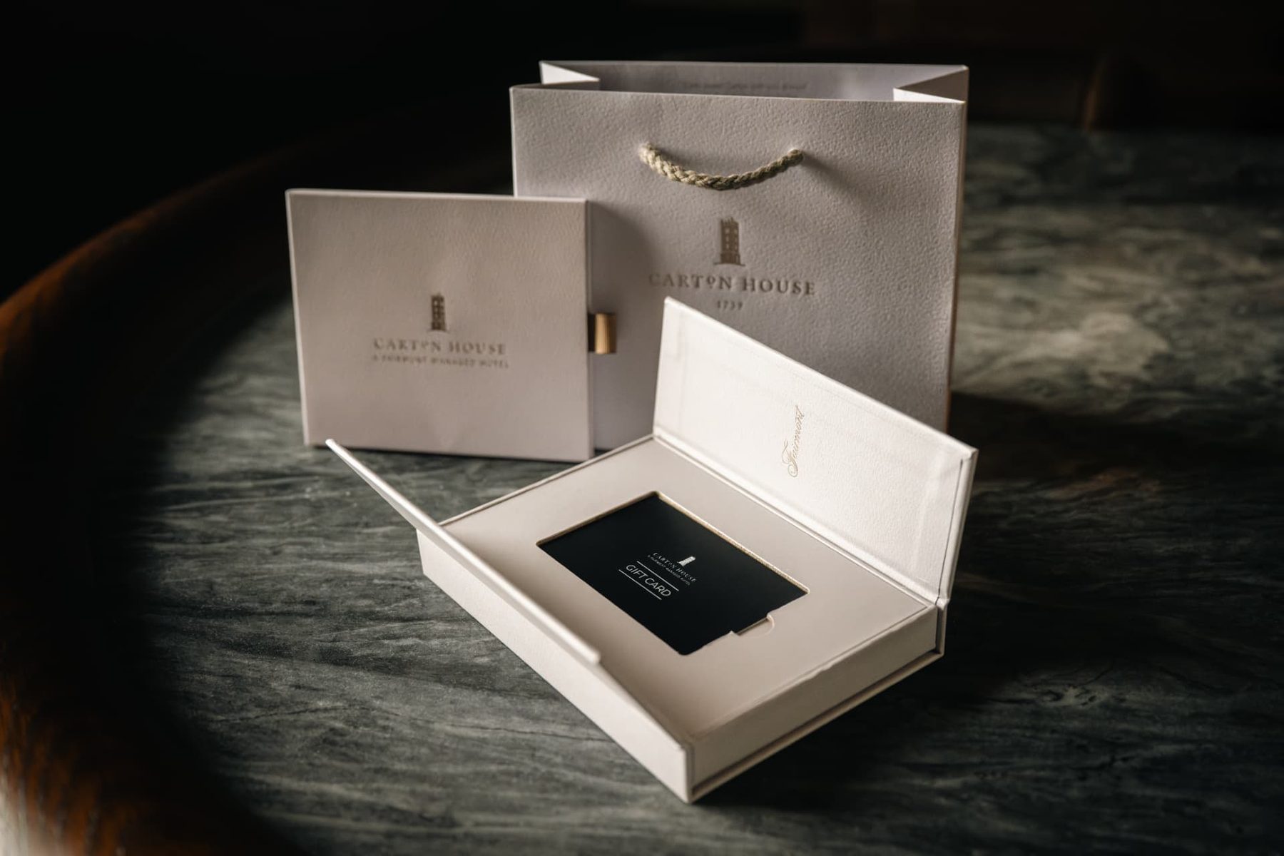 Two matt off white gift boxes and a matching rope handle paper bag with gold foil logos.