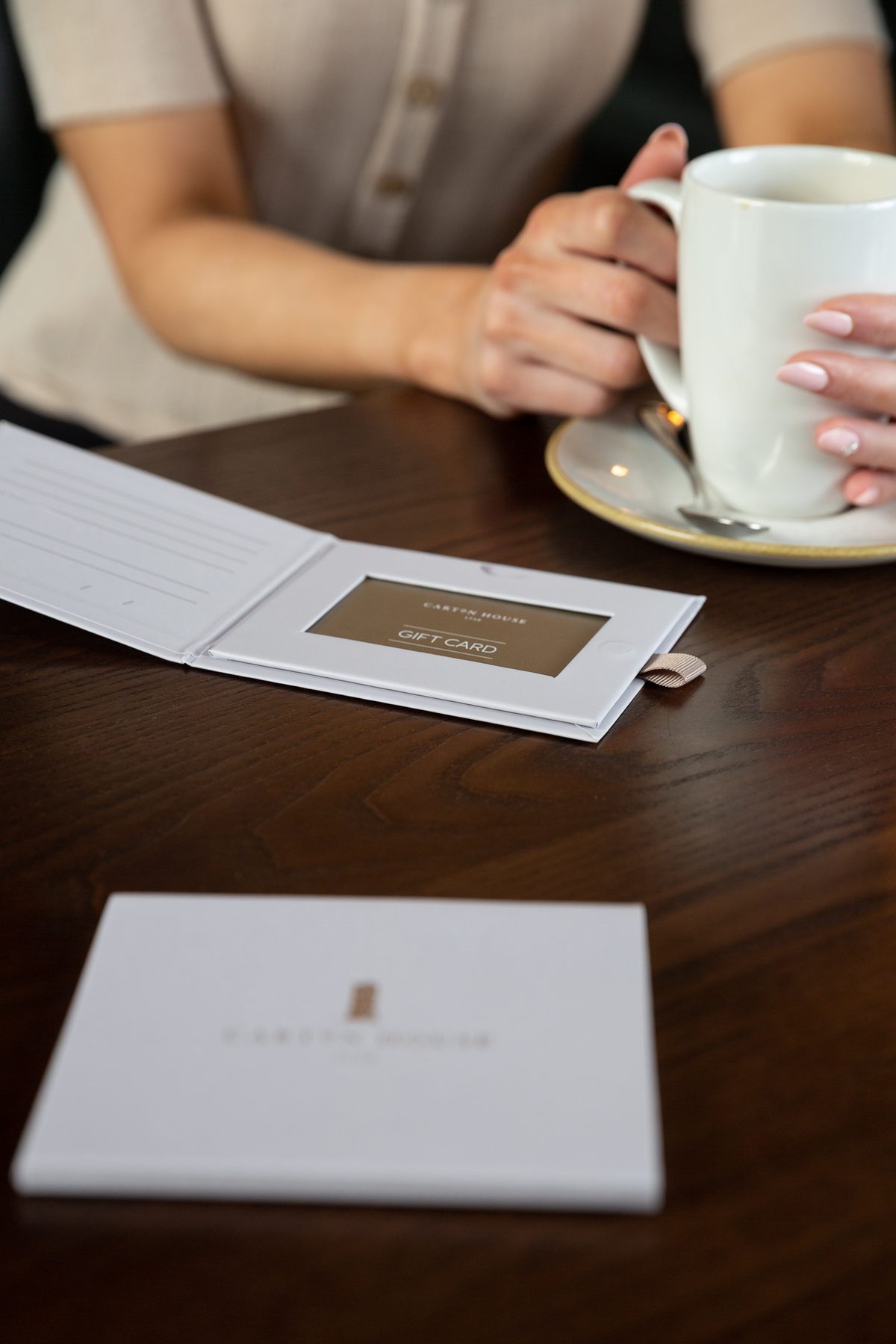 White, rigid, landscape gift card wallet. Gold foil branding. A light brown gift card shows through a die cut window and features a ribbon pull.